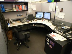 Desk in a cubby space with papers, two desktop computer screens and a chair